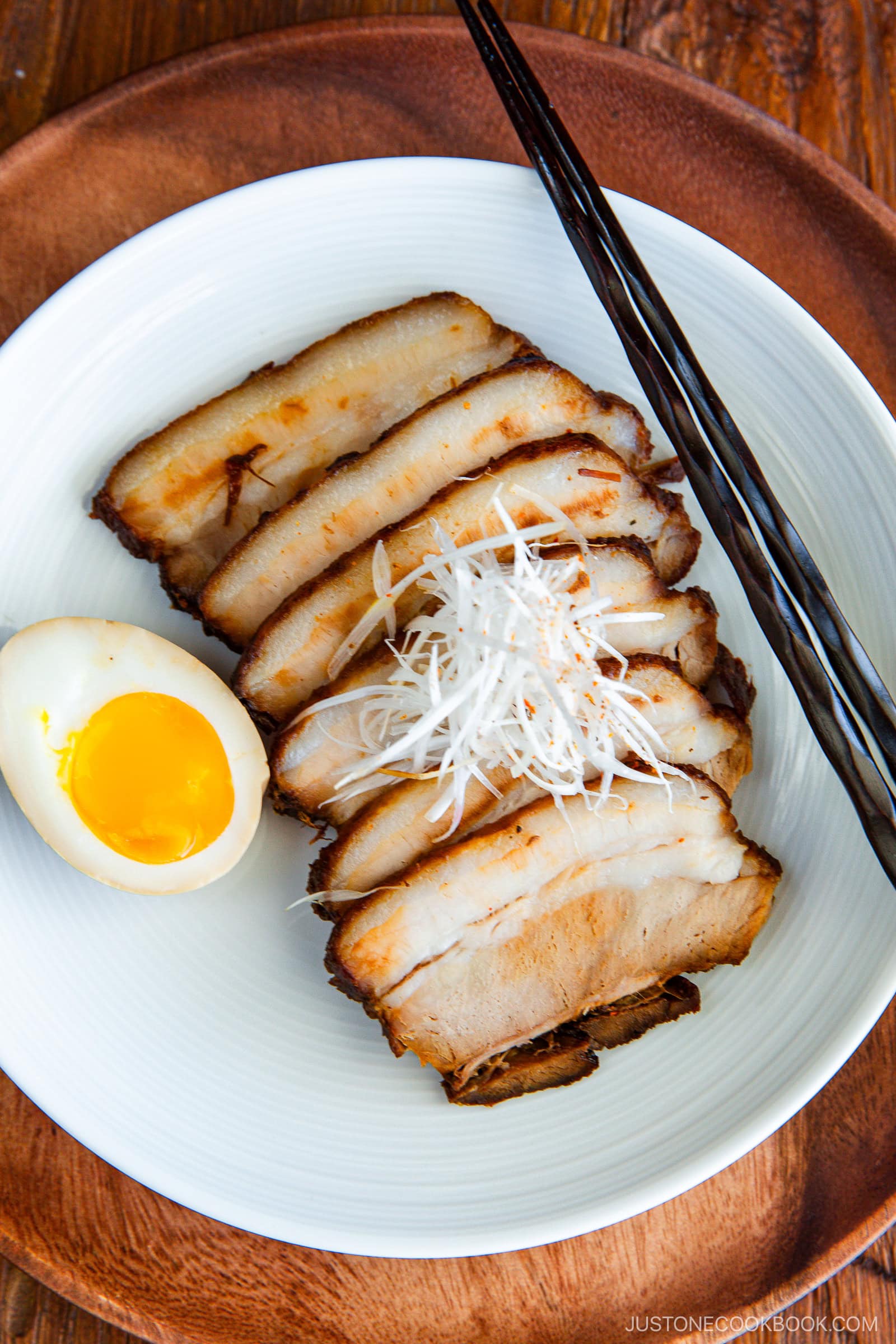 A white plate with sliced chashu pork belly, a soft-boiled egg with a runny yolk, and shredded white scallions, served with black chopsticks on the side.