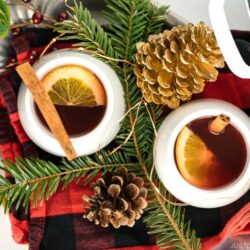 White cups containing hot mulled cider garnished with orange slice and a cinnamon stick.