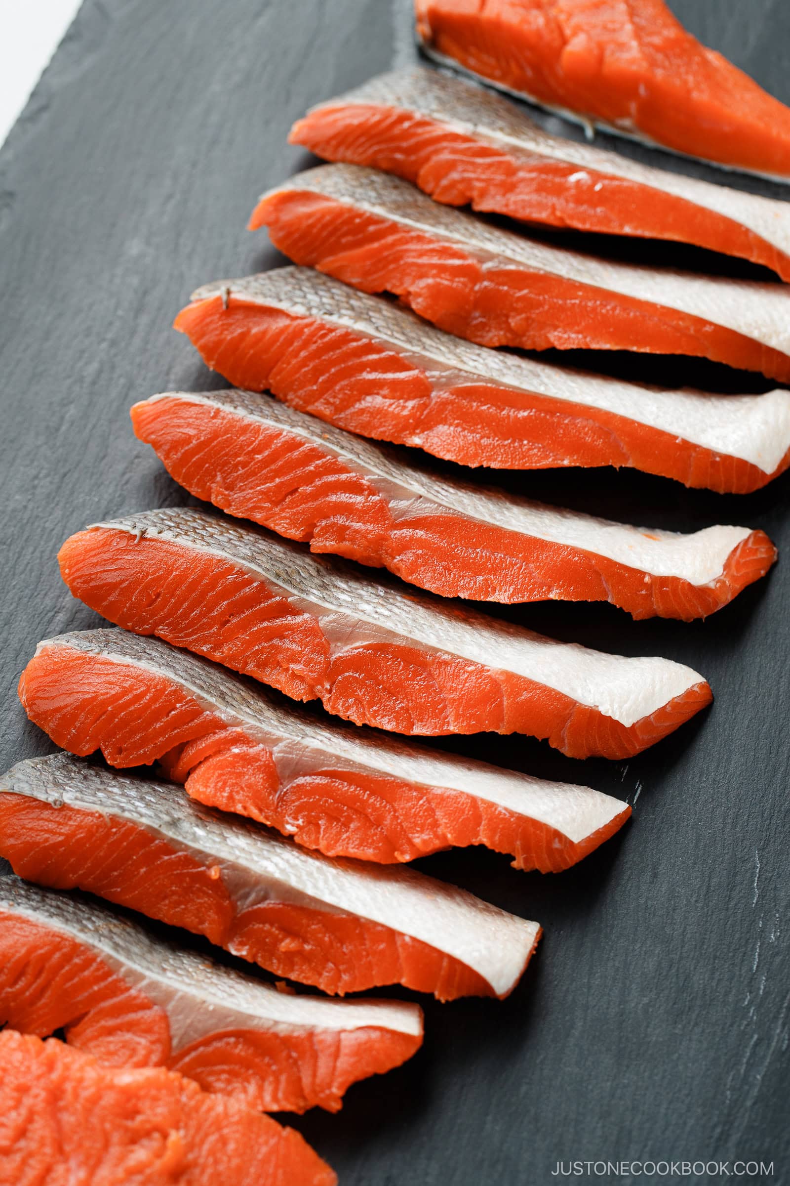 Sliced raw salmon with silver skin and vibrant orange flesh, arranged neatly in a row on a dark slate board, showcasing how to cut salmon into Javanese style fillets.
