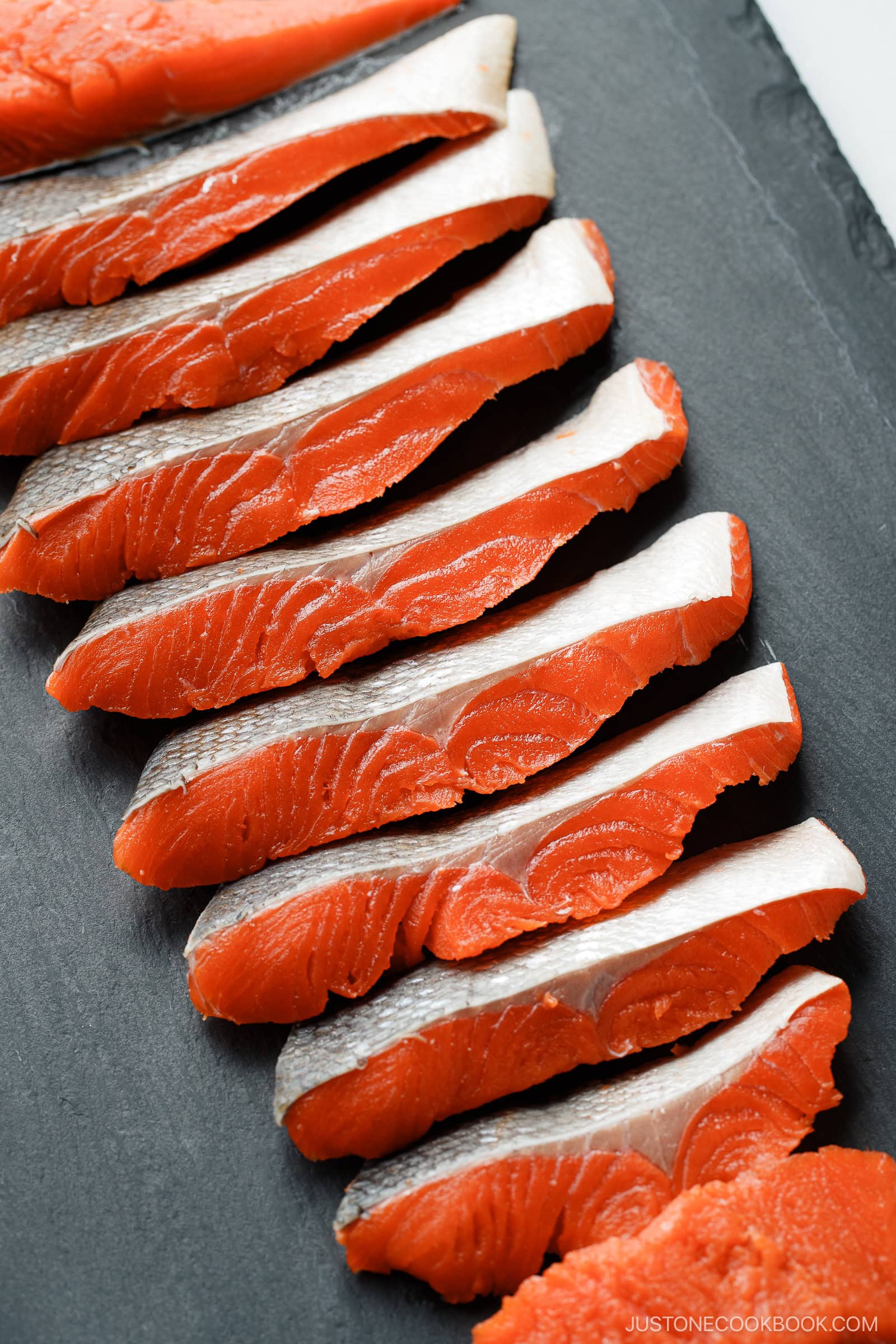 Slices of raw salmon, expertly prepared following how to cut salmon into Javanese style fillets, showcase vibrant orange flesh and silvery skin, neatly arranged in a row on a dark slate surface.