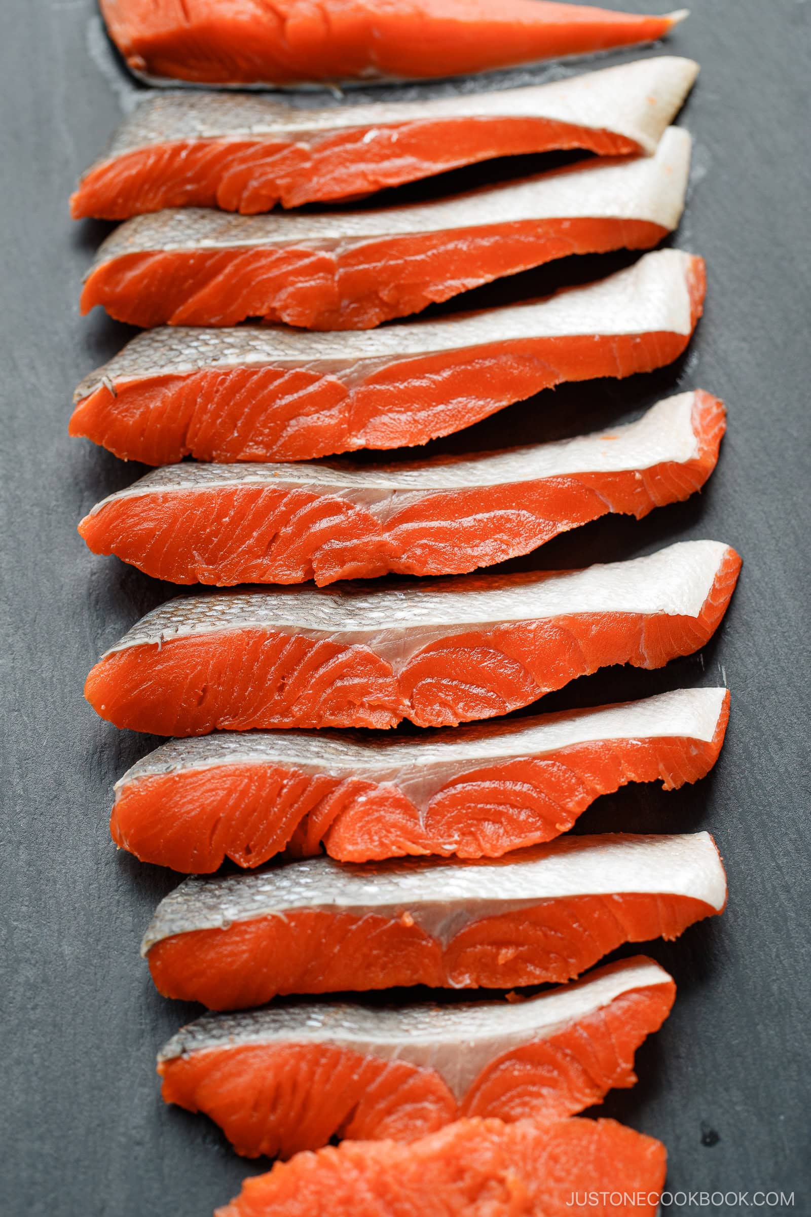 Several raw salmon fillets with silver skin and bright orange flesh are neatly arranged in a row on a dark surface, showcasing how to cut salmon into Javanese style fillets.