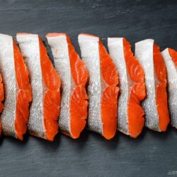 Slices of raw salmon with silver skin and bright orange flesh are neatly arranged in a row on a dark slate surface, showcasing how to cut salmon into Javanese style fillets.