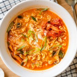 A white ramen bowl containing Korean ramen (ramyeon), topped with soft-cooked egg, kimchi, and green onions.
