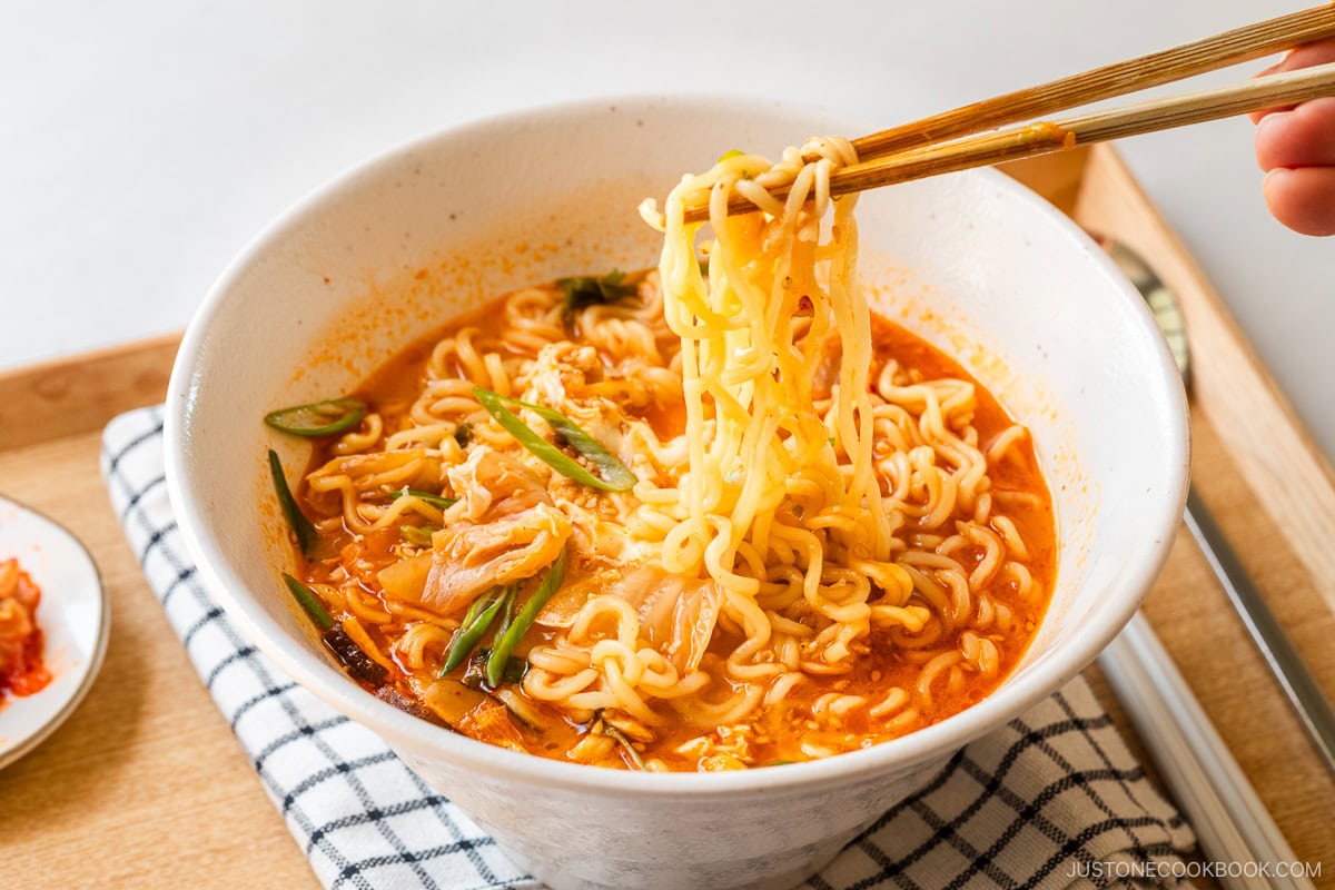 A white ramen bowl containing Korean ramen (ramyeon), topped with soft-cooked egg, kimchi, and green onions.