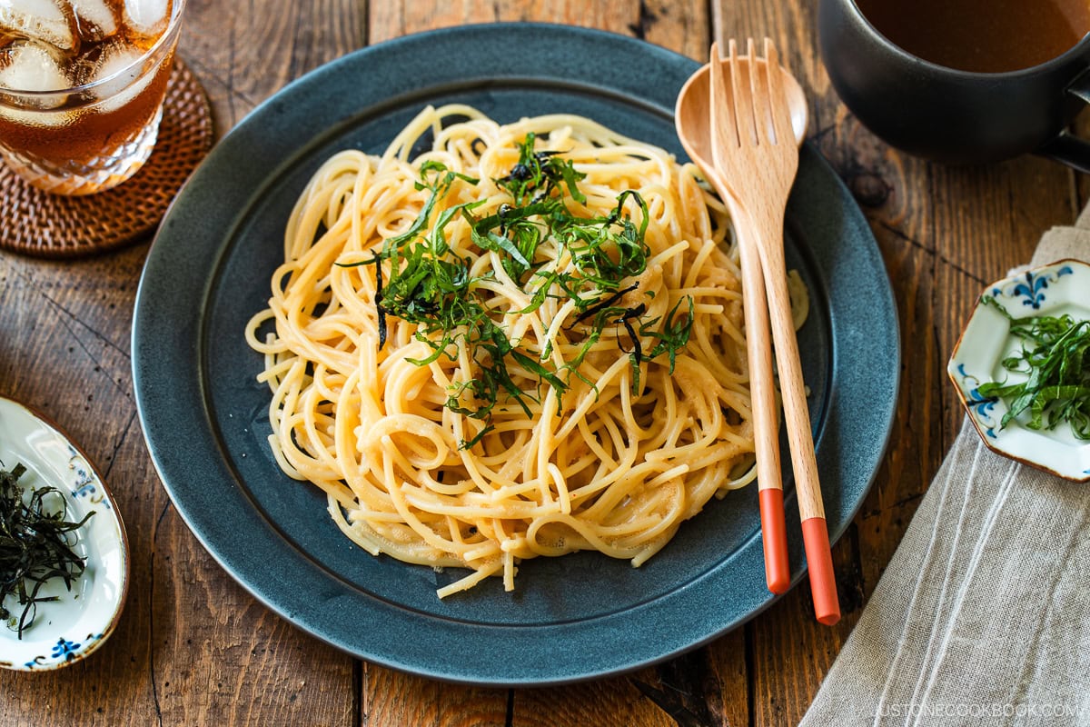 A black round plate containing metaiko pasta - Japanese spaghetti with spicy salted cod roe topped with julienned shiso and shredded nori seaweed.