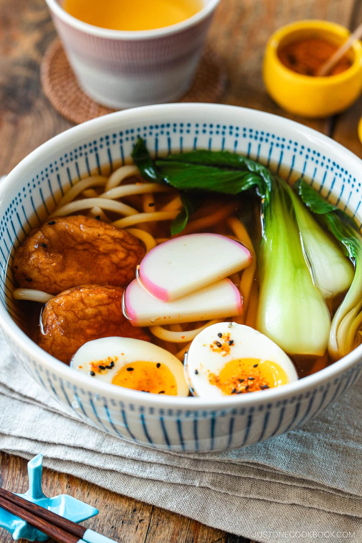 A large blue and white bowl containing udon noodle soup topped with fish cake, bok choy, fish cakes, and jammy soft boiled eggs in a savory dashi broth.