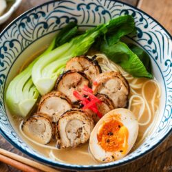 Blue and white bowls containing ramen with soup topped with seared sliced chicken chashu, soft boiled egg, and blanched bok choy, and garnished with pickled ginger.