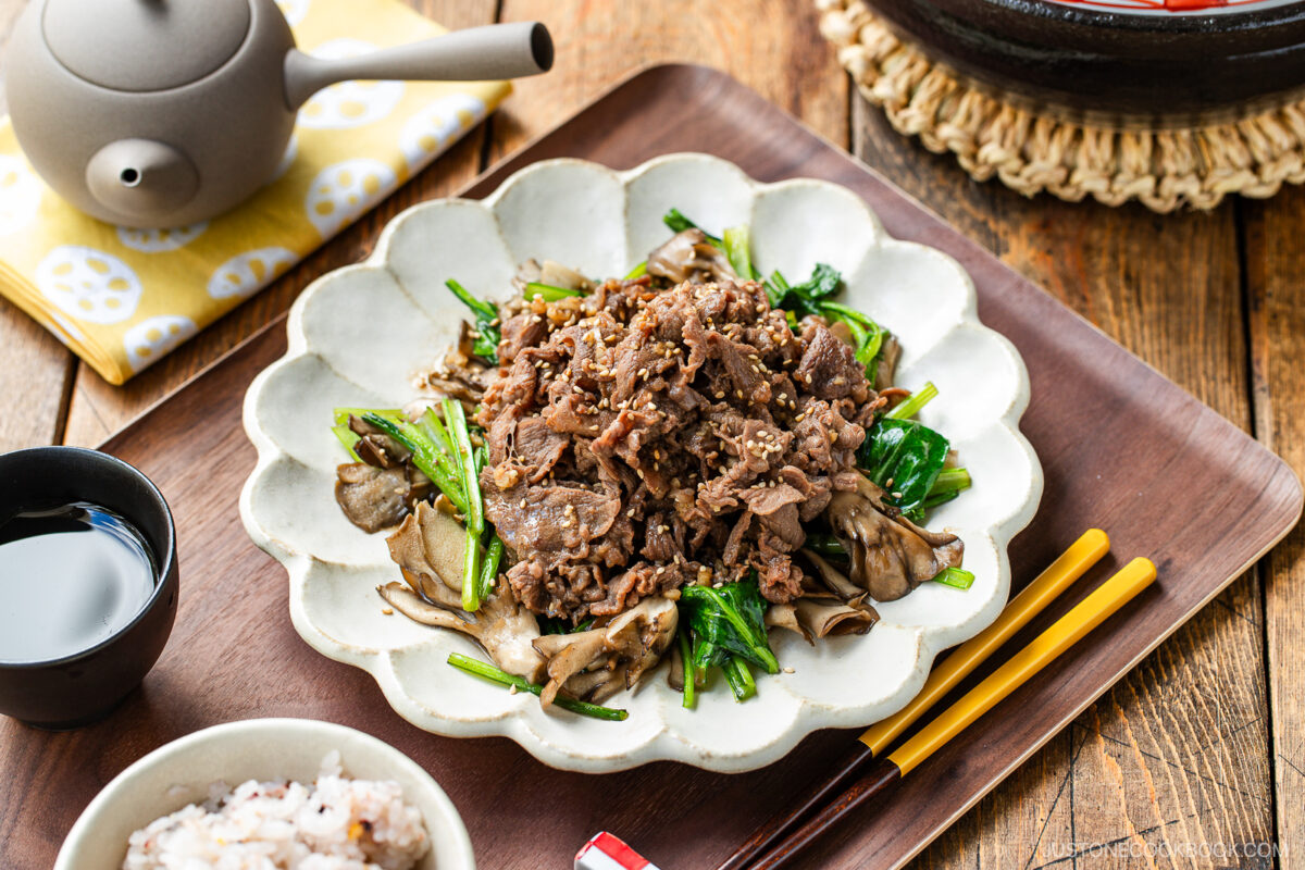 A white scalloped plate containing stir-fried Butter Ponzu beef with komatsuna green and maitake mushrooms.