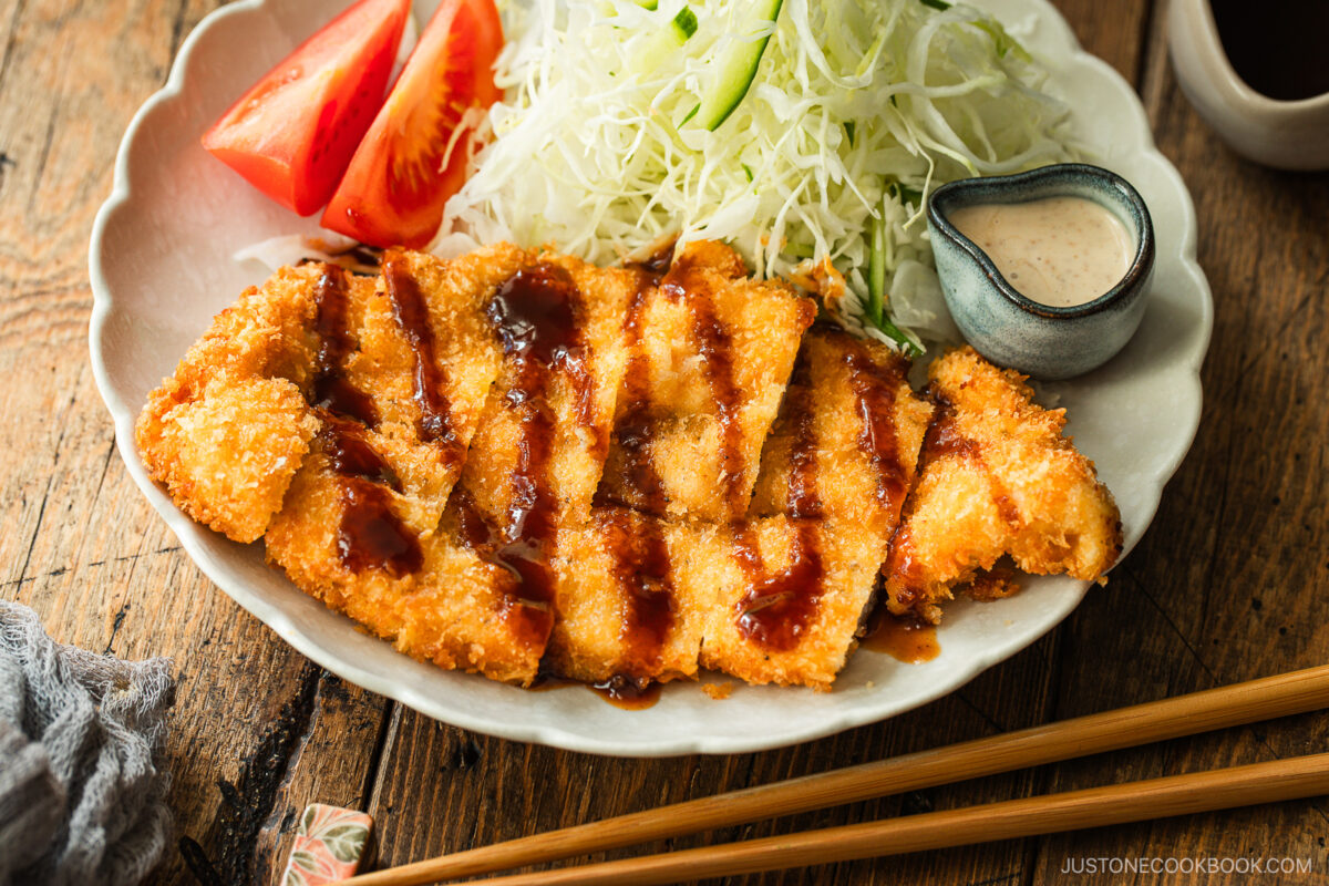 A plate of crispy breaded chicken cutlets drizzled with sauce, served with shredded cabbage, tomato wedges, chopsticks, and a small dish of creamy dressing on a wooden table.