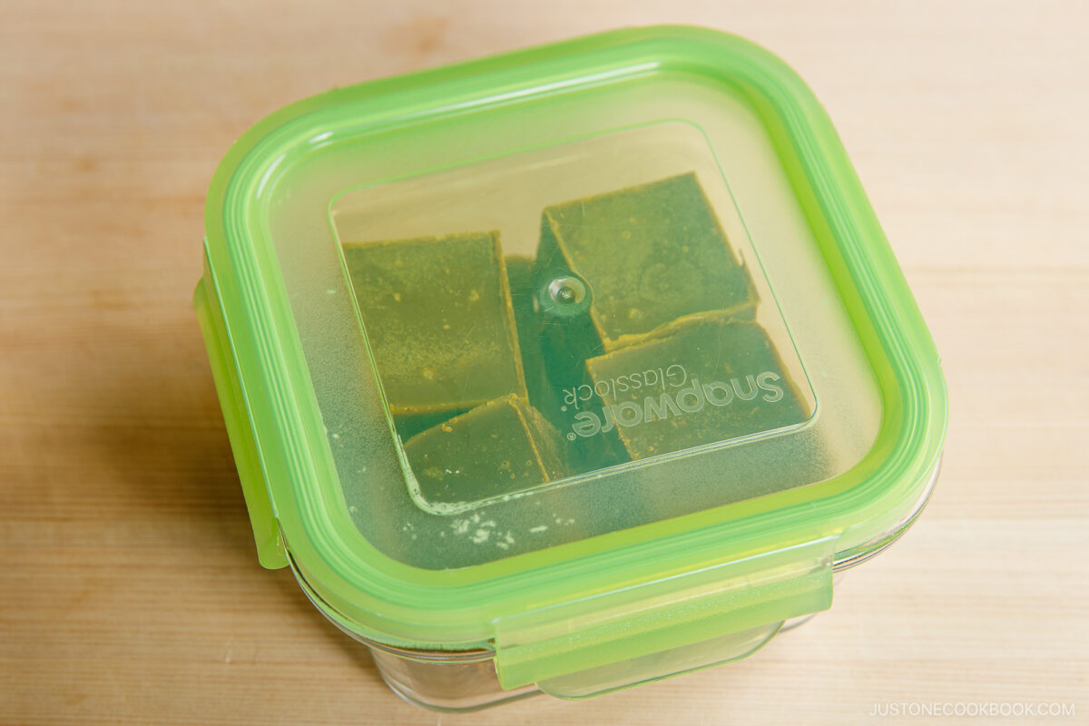 A green-lidded glass container holds several green matcha cubes, visible through the transparent lid, placed on a light wooden surface.
