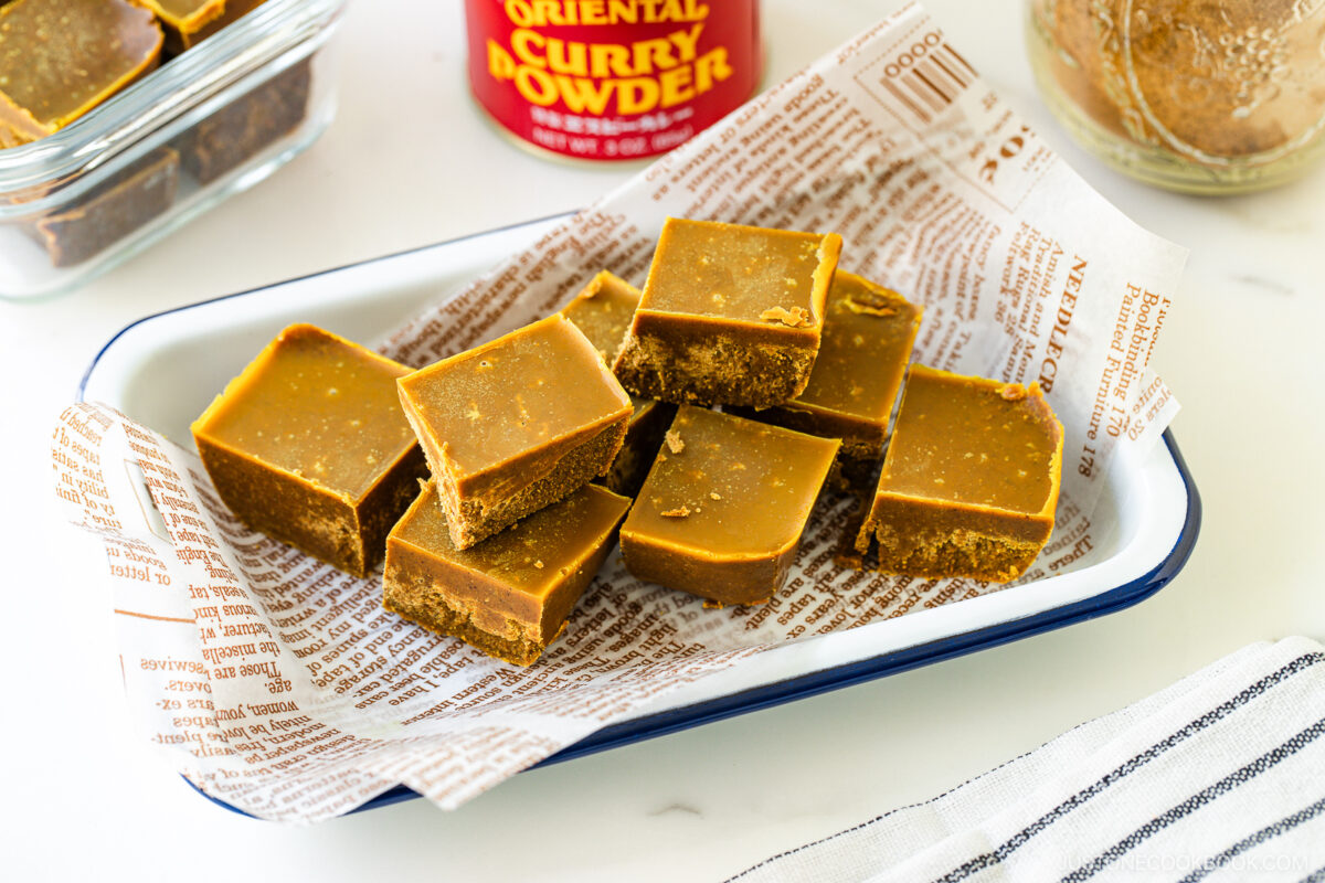 A white enamel tray lined with printed paper holds several pieces of golden brown fudge. A jar labeled Oriental Curry Powder and a glass container of more fudge are in the background.