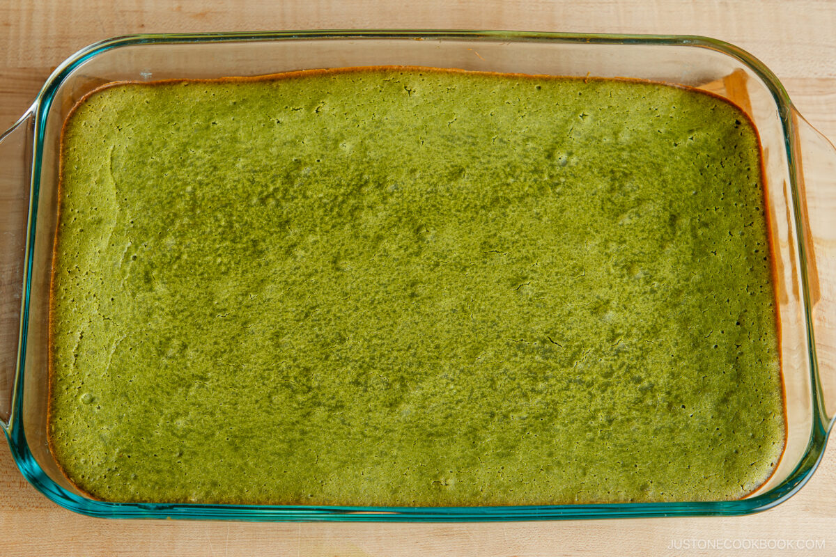 A rectangular glass baking dish filled with a freshly baked green matcha cake, with a slightly browned edge resting on a wooden surface.