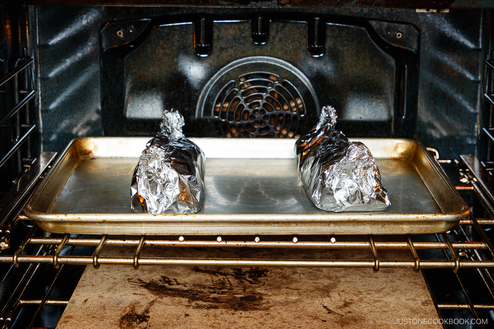 Two foil-wrapped packets sit on a baking sheet inside an oven, mid-baking. The oven racks and fan are visible in the background.