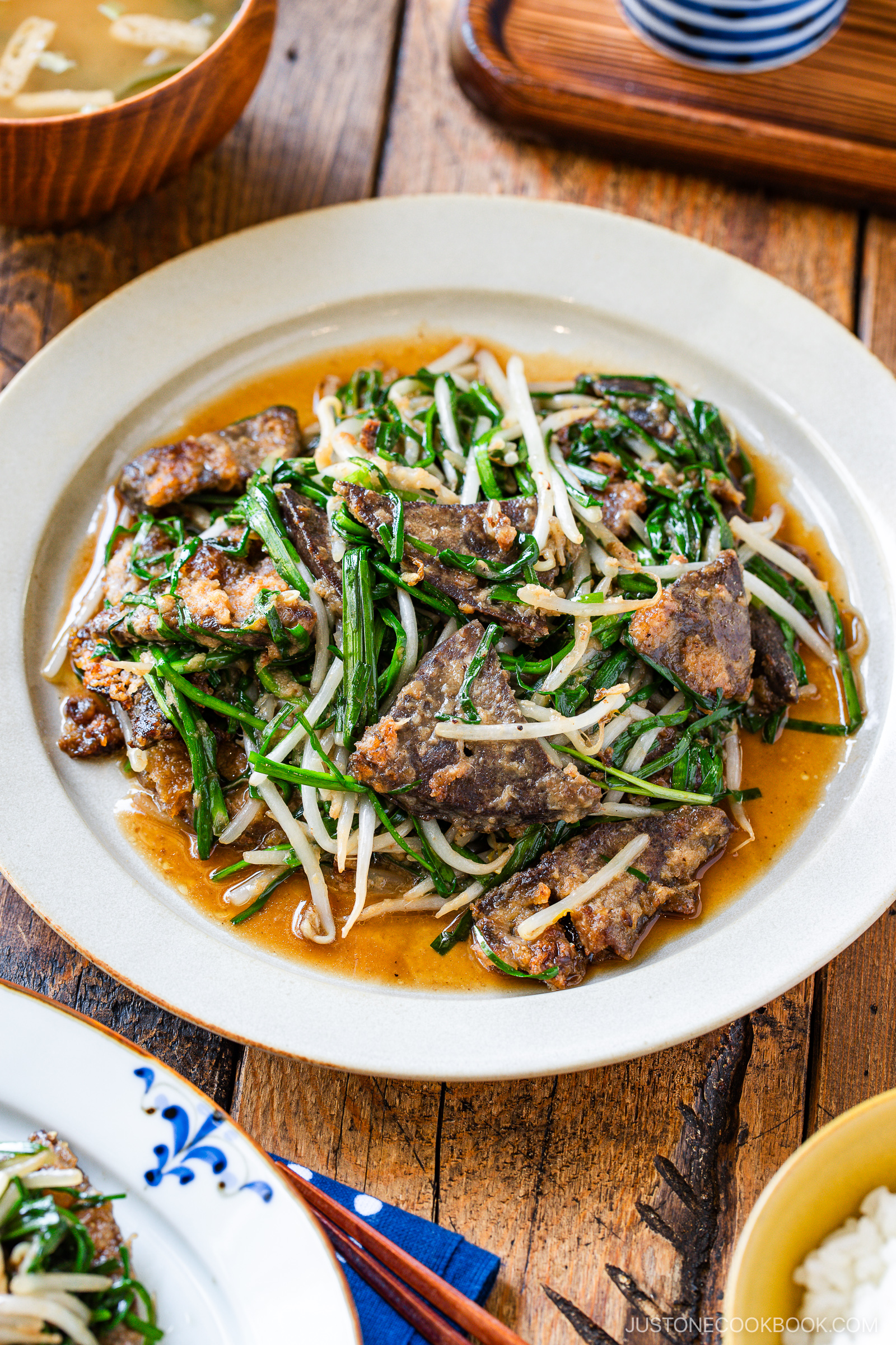 A plate of stir-fried beef liver with bean sprouts and green vegetables in a savory brown sauce, served on a white dish on a wooden table. Miso soup and rice are partially visible in the background.
