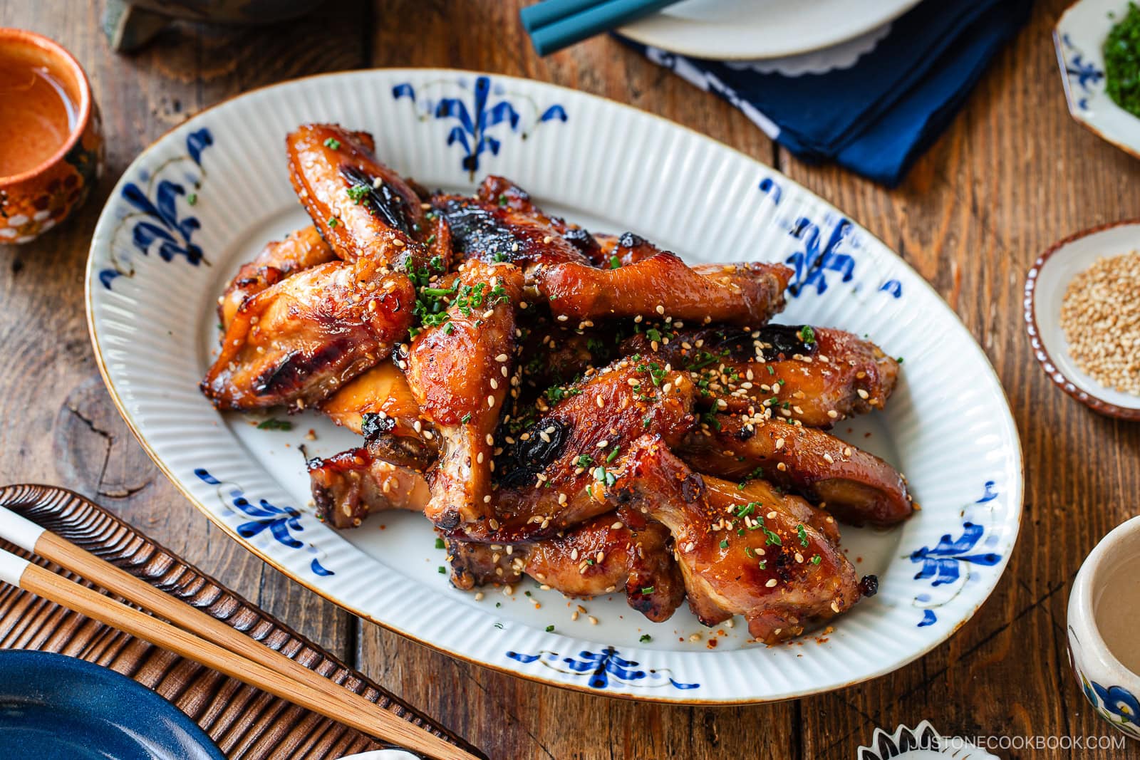 A plate of glazed chicken drumsticks garnished with sesame seeds and chopped herbs, served on a white and blue decorative dish atop a wooden table set with chopsticks and small bowls.