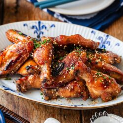 A platter of glazed chicken wings garnished with chopped green herbs and sesame seeds, served on a decorative oval plate on a wooden table.