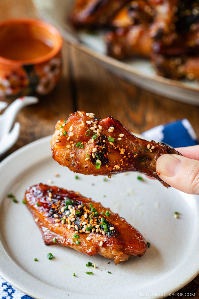 A hand holds a glazed chicken wing garnished with sesame seeds and chopped herbs over a white plate, which has another similar wing resting on it. A bowl of sauce and more wings are visible in the background.