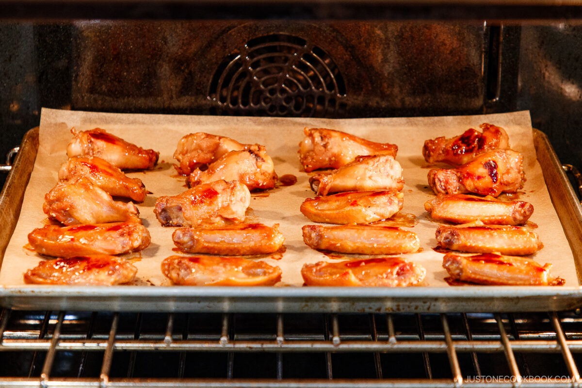 Chicken wings arranged on a baking sheet with parchment paper, cooking inside an oven. The wings appear to be partly cooked, with a slight glaze visible on some pieces.