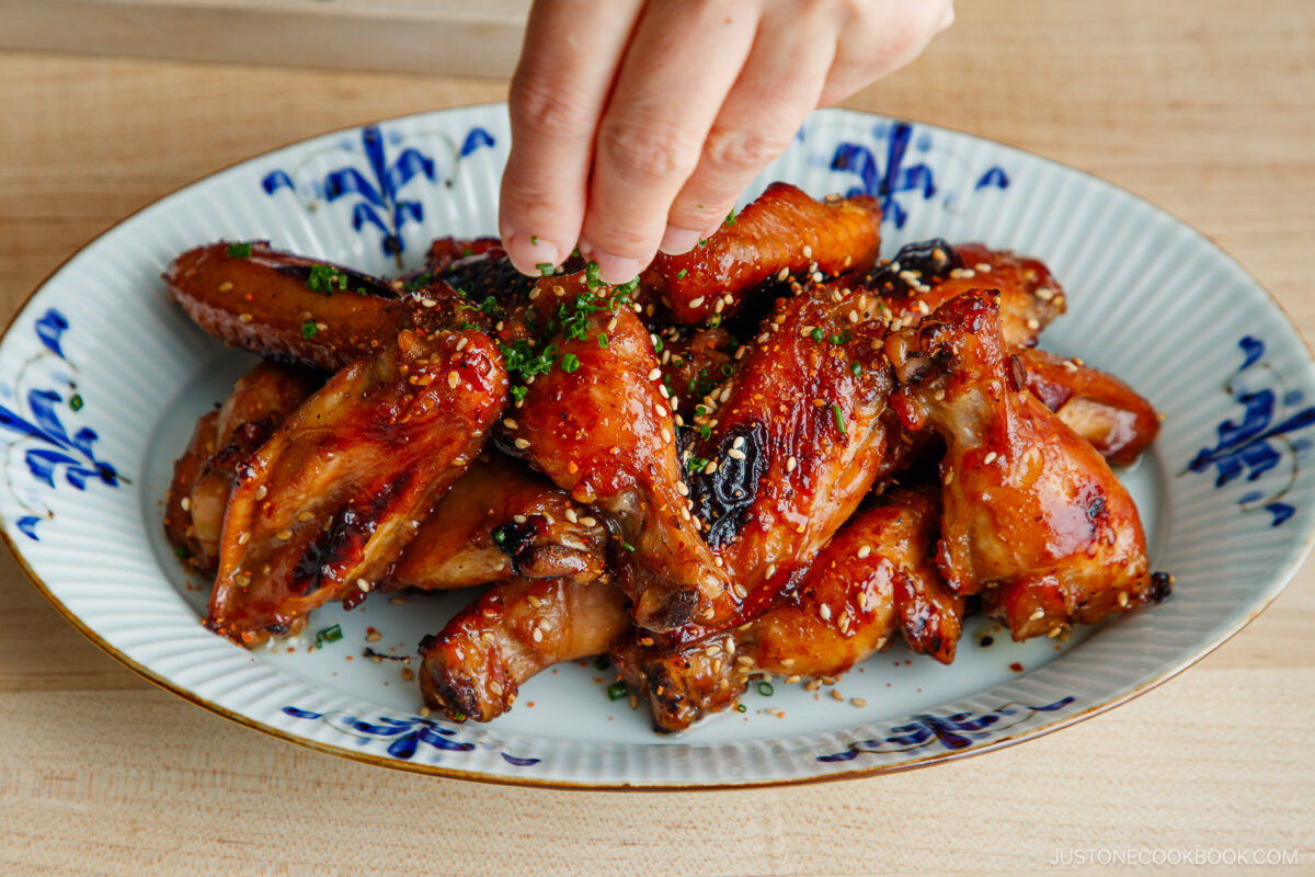 A hand sprinkles chopped herbs onto a plate of glazed chicken wings garnished with sesame seeds, served on a white dish with blue patterns, placed on a wooden table.