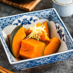 A decorative blue-and-white bowl filled with simmered kabocha squash pieces, garnished with thinly sliced ginger. Chopsticks and small dishes are placed beside the bowl on a dark surface.