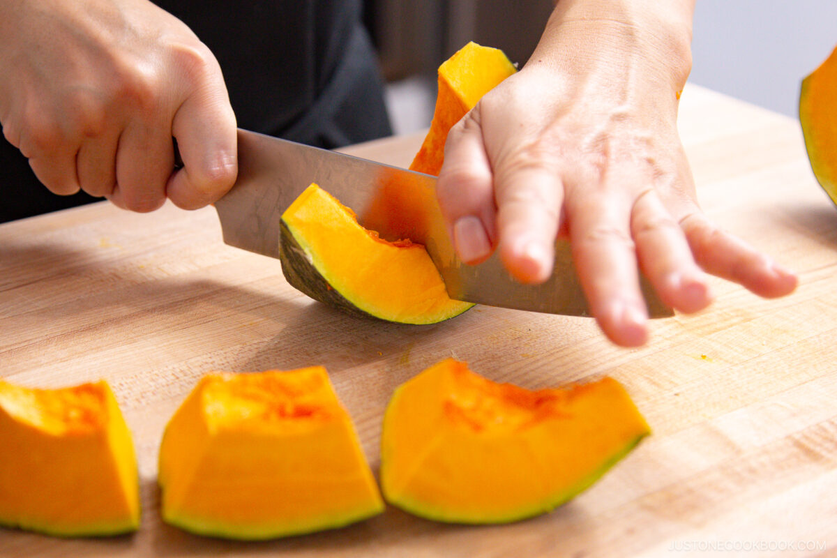 A person’s hands slicing a bright orange kabocha squash on a wooden cutting board, with several squash wedges already cut and arranged in front.