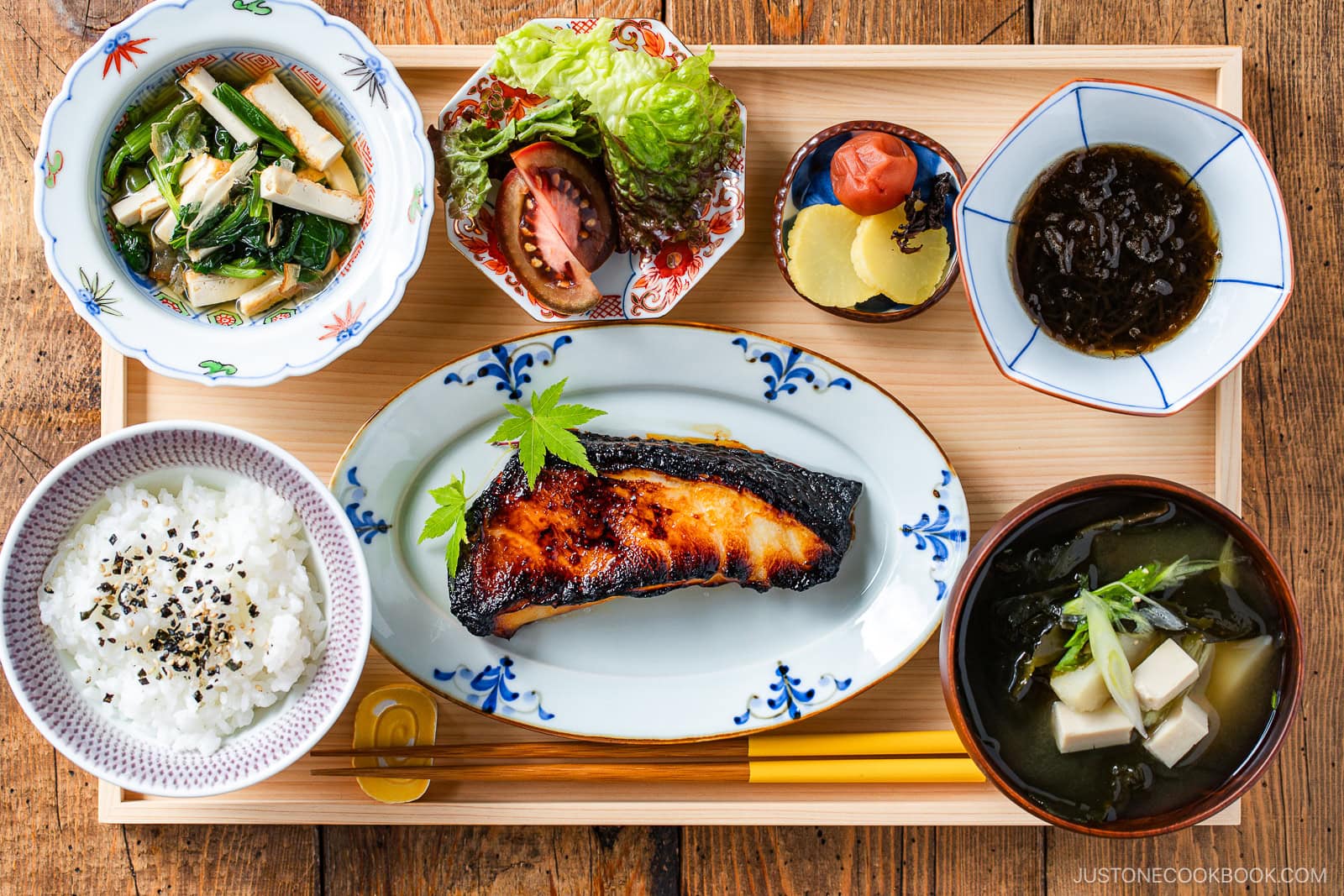 A traditional Japanese meal set on a wooden tray, featuring grilled fish, rice, miso soup, leafy greens, pickles, seaweed, and fresh vegetables in small bowls. Chopsticks and a spoon are placed at the front.