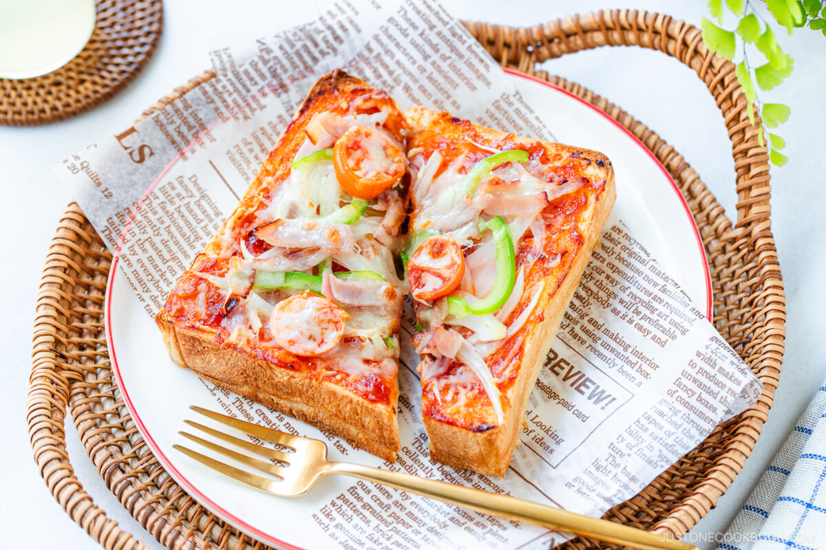 A slice of thick toast topped with melted cheese, sliced onions, cherry tomatoes, green peppers, and sausage, served on a plate lined with printed paper, with a gold fork on the side.