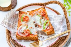 A slice of toast topped with melted cheese, cherry tomatoes, onions, and green peppers on a decorative plate, served with a gold fork on a wicker tray lined with newspaper-style parchment.