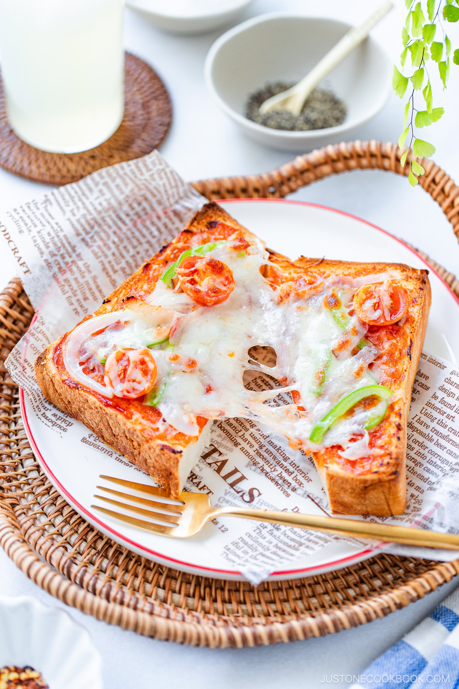 A slice of toast topped with melted cheese, cherry tomatoes, green bell pepper, and onion sits on a plate lined with newspaper-style paper, with a golden fork beside it. In the background are small bowls with condiments.