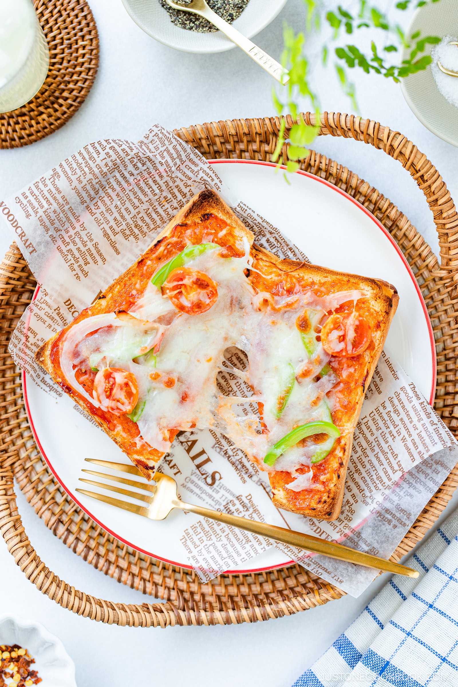 A slice of toast topped with melted cheese, cherry tomatoes, green peppers, and onions sits on a plate lined with decorative paper, placed on a woven tray with a gold fork beside it.