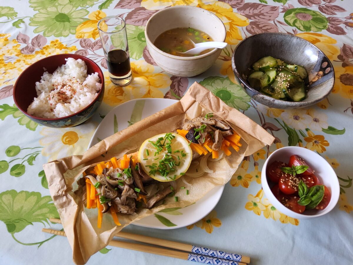 A Japanese meal with rice, miso soup, cucumber salad, tomato salad, and a main dish of fish, mushrooms, carrots, and lemon on parchment paper, all served on a floral tablecloth with chopsticks and a drink.