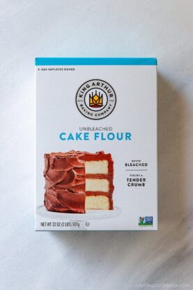 A box of King Arthur Baking Company Unbleached Cake Flour, featuring an image of a layered cake with chocolate frosting. The box is 32 oz and emphasizes tender crumb and never bleached.