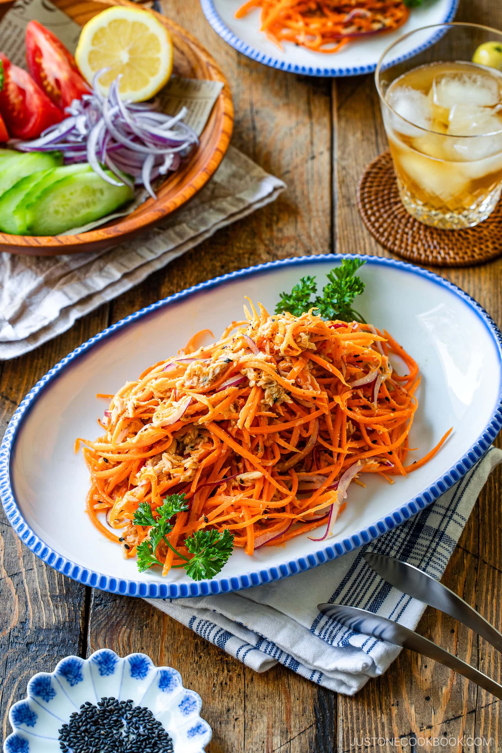 A plate of shredded carrot salad garnished with parsley sits on a rustic table, accompanied by a glass of iced drink, a dish of black sesame seeds, and a plate of sliced cucumber, tomato, lemon, and red onion.