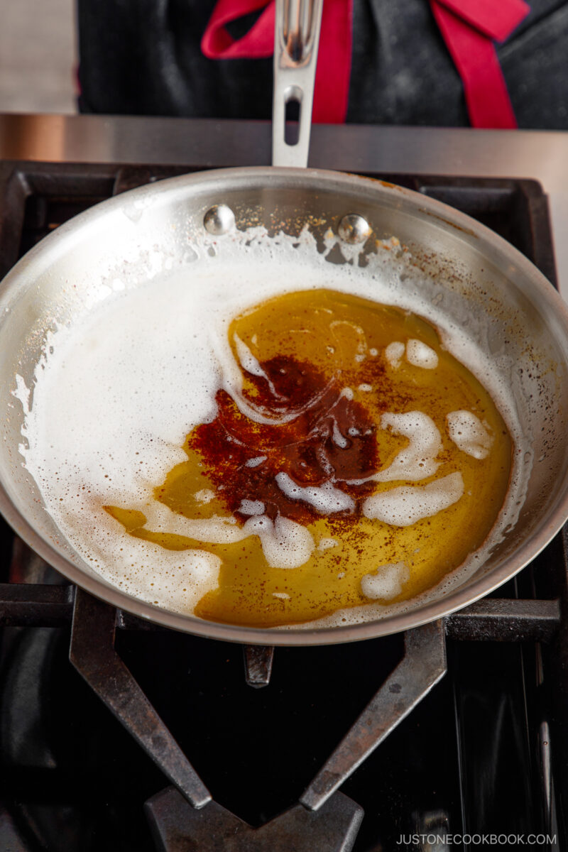 A stainless steel pan on a stovetop contains melted butter browning, with golden brown bits and white foam. A person in dark clothing with a red tie is partially visible in the background.