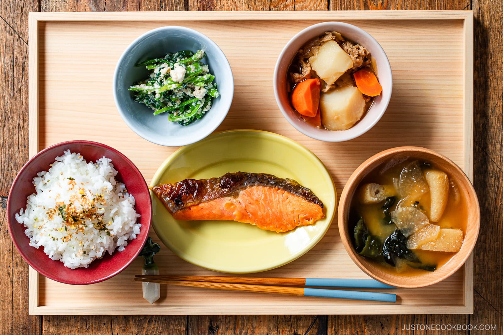 A wooden tray with a Japanese meal: grilled salmon, miso soup, white rice, spinach with sesame, and simmered vegetables with meat, each in separate bowls on a wooden table. Chopsticks rest beside the tray.