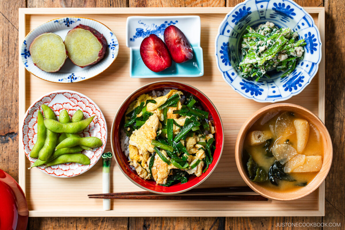 A Japanese meal on a wooden tray featuring a bowl of rice with vegetables and egg, miso soup, edamame, sweet potato slices, plums, and a green vegetable side dish, with chopsticks and a small sauce dish.