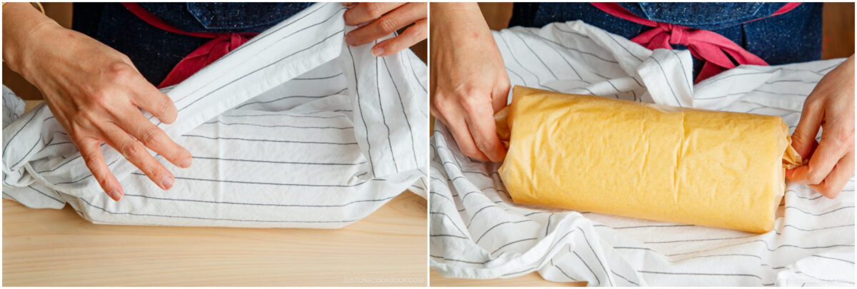 A person in a blue apron with a red tie wraps a rolled cake in parchment paper using a striped kitchen towel on a wooden surface.