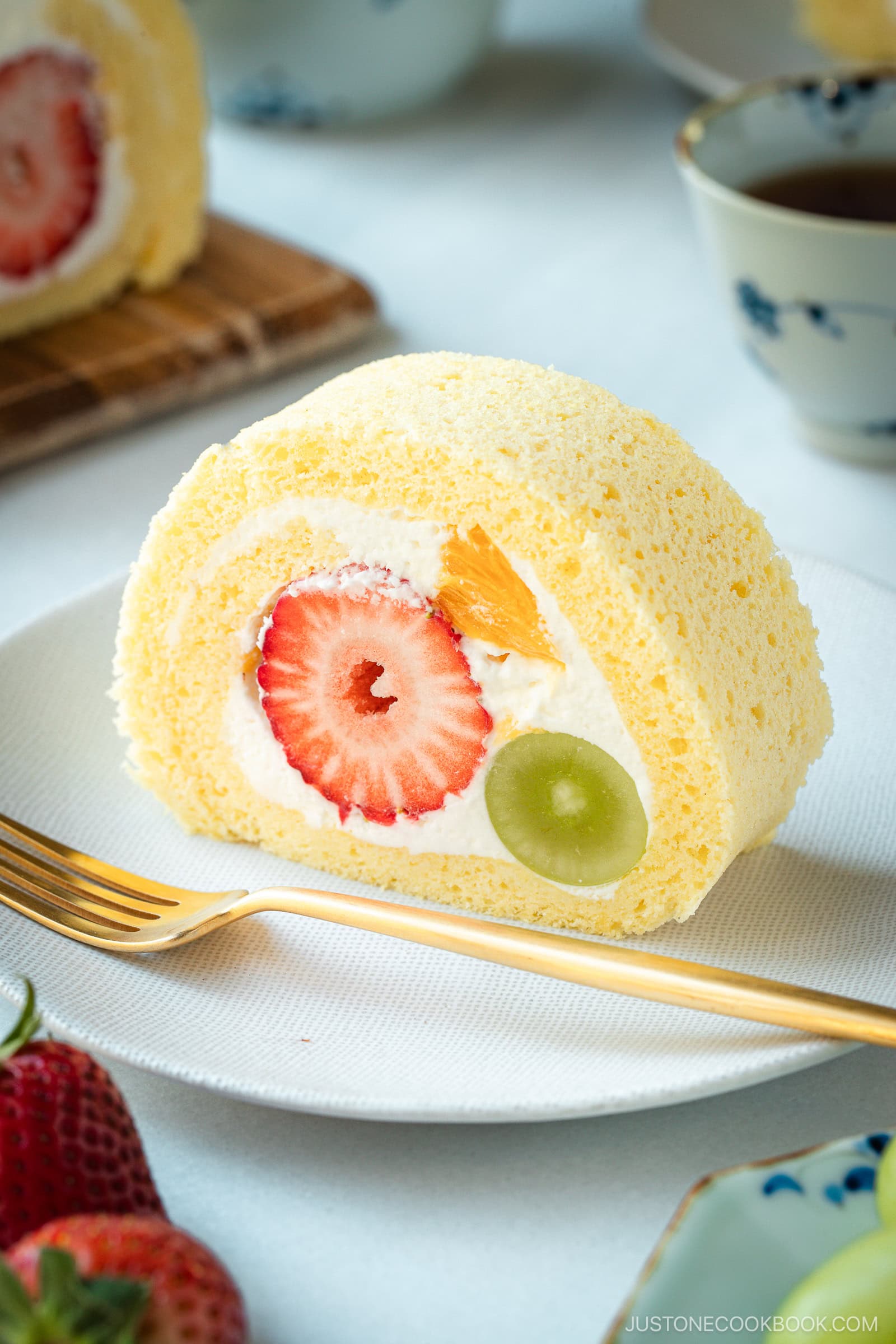 A slice of Japanese fruit roll cake filled with whipped cream, a whole strawberry, a green grape, and a piece of orange, served on a white plate with a gold fork and fresh strawberries on the side.