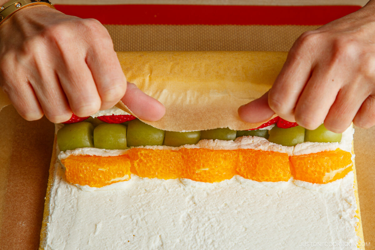Hands carefully rolling a fruit-filled cake roll with layers of orange slices, green grapes, and strawberries on a sheet of whipped cream-covered sponge cake.