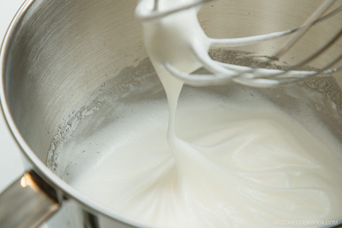 A metal mixing bowl with glossy, whipped egg whites forming soft peaks, and a whisk attachment covered in the mixture.