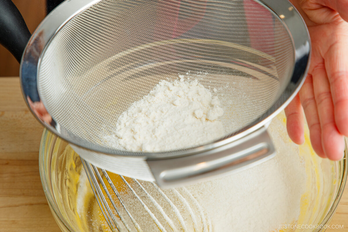 A hand holds a metal sifter filled with flour over a glass mixing bowl containing yellow batter, ready to sift the flour into the mixture.