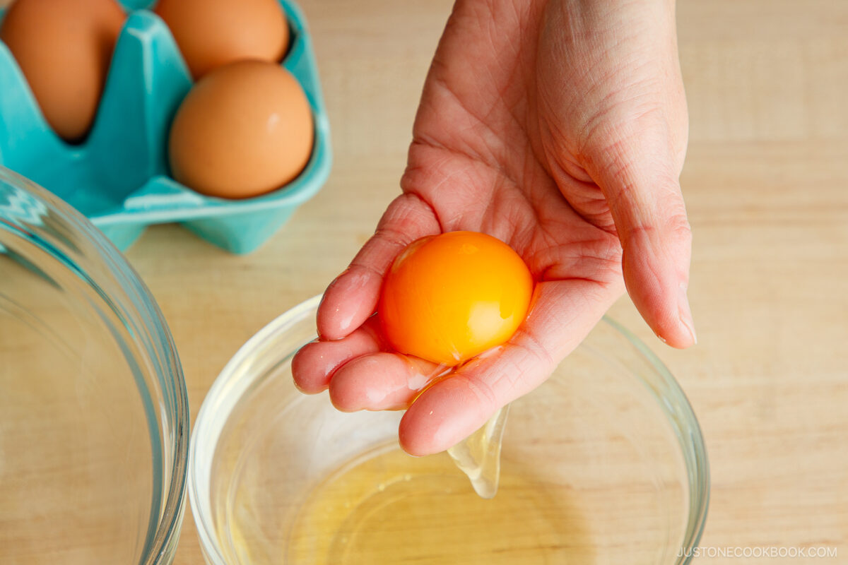 A hand holding a raw egg yolk over a glass bowl, with egg white in the bowl below and brown eggs in a blue carton in the background.