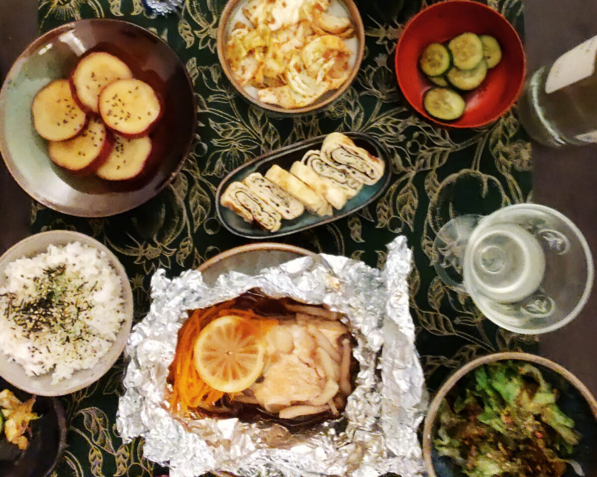 A Japanese meal with rice, sweet potatoes, pickled cucumbers, tamagoyaki, salad, foil-baked fish topped with lemon and carrots, and cabbage, served on a floral-patterned tablecloth with a glass of wine.
