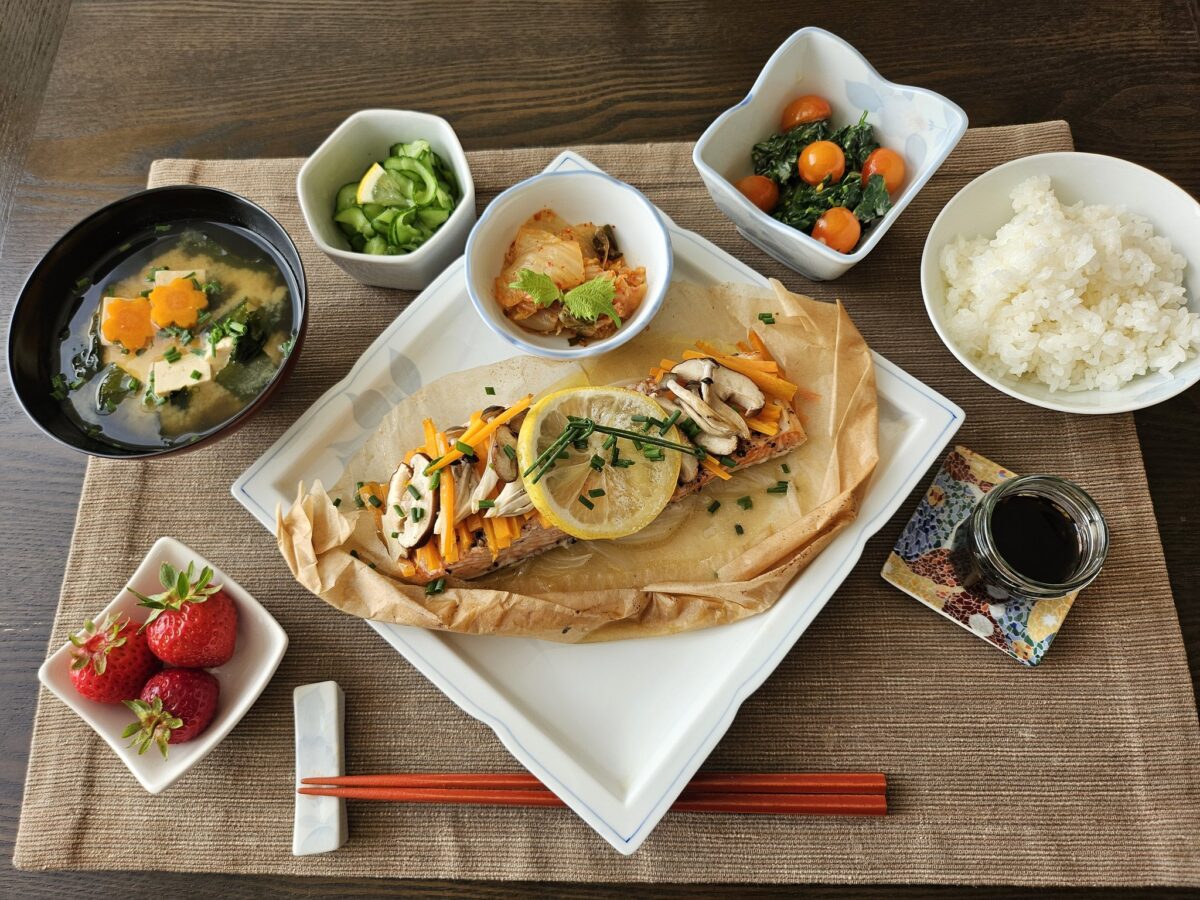A Japanese meal set on a brown placemat, featuring rice, miso soup, cooked fish with vegetables, pickled cucumbers, cherry tomato salad, kimchi, fresh strawberries, soy sauce, and wooden chopsticks.