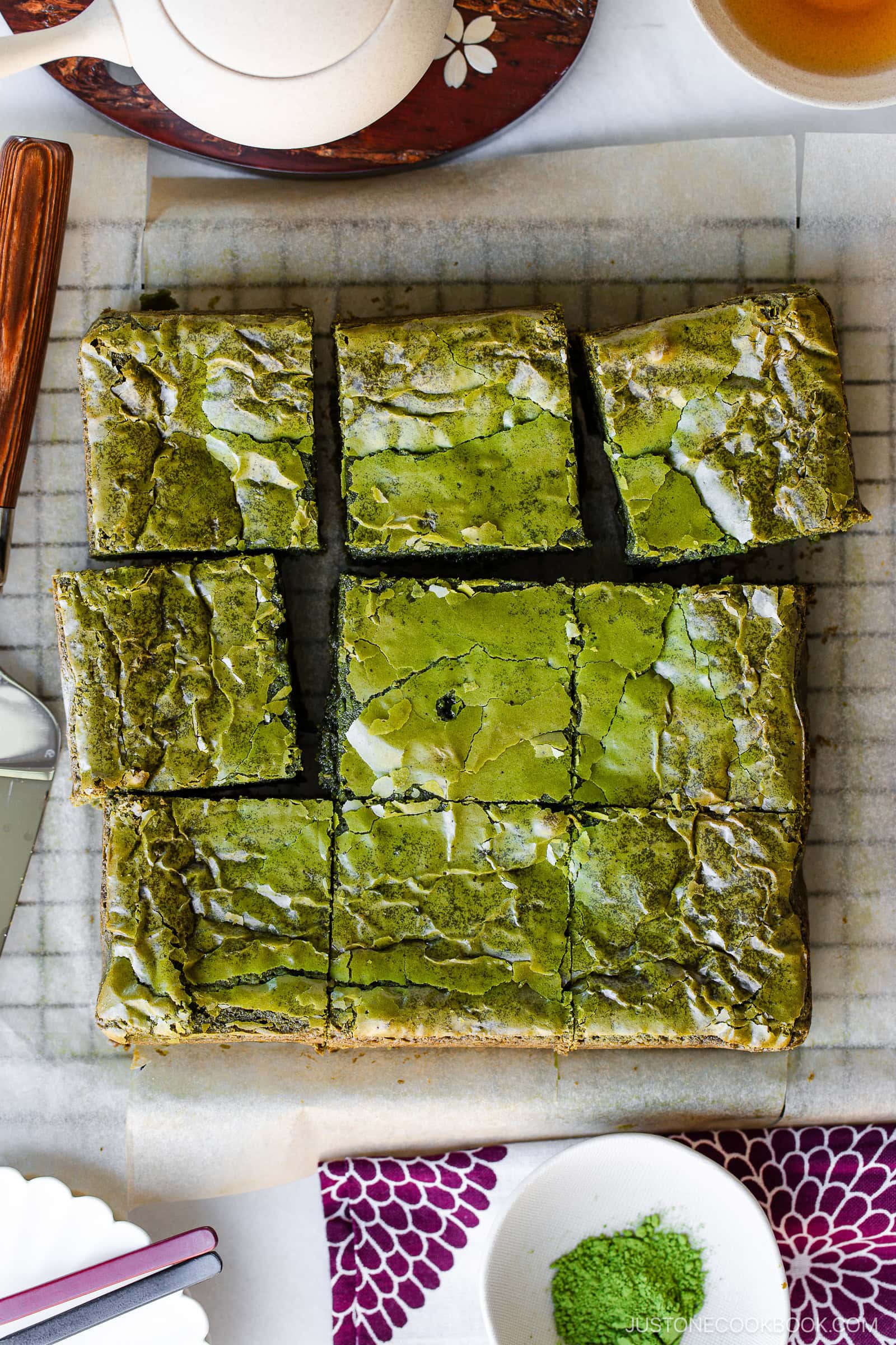 Nine square pieces of green matcha brownies with a shiny, slightly cracked surface are arranged on parchment paper. A teapot, tea cup, utensils, and a bowl of matcha powder are nearby.