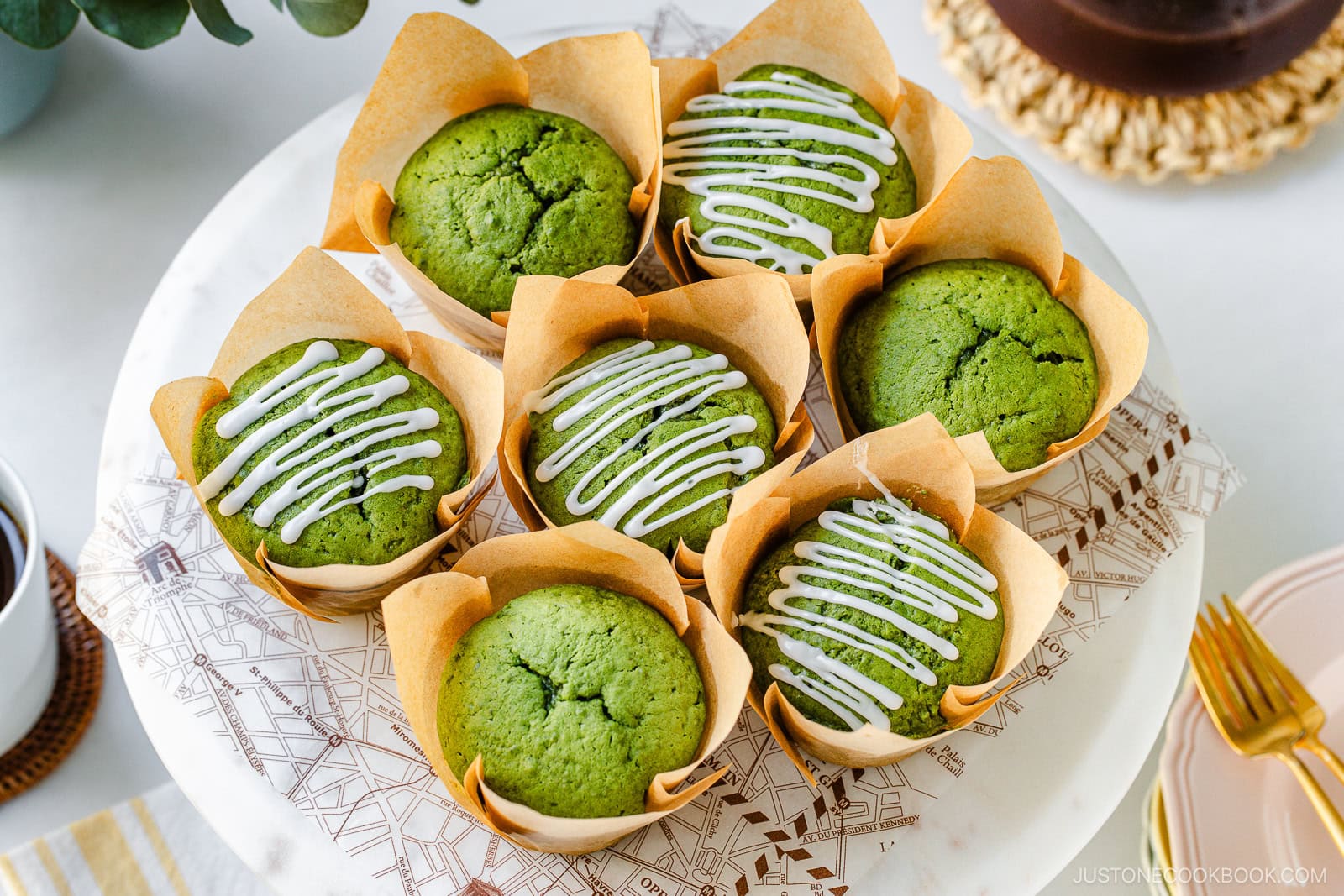 Seven matcha muffins in brown paper wrappers are arranged on a white plate. Four muffins have white icing drizzled on top, while the other three are plain. The scene is bright and inviting, with a hint of a coffee cup and utensils nearby.