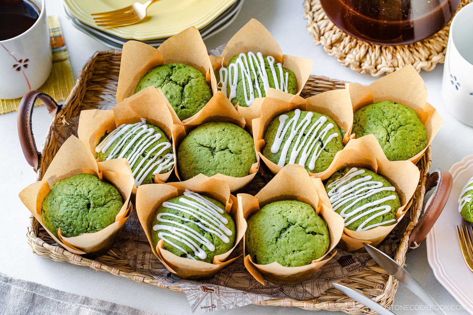 A wicker tray holds nine green muffins in brown parchment wrappers. Some muffins are drizzled with white icing, while others are plain. Plates, coffee cups, and a coffee pot surround the tray on a white table.