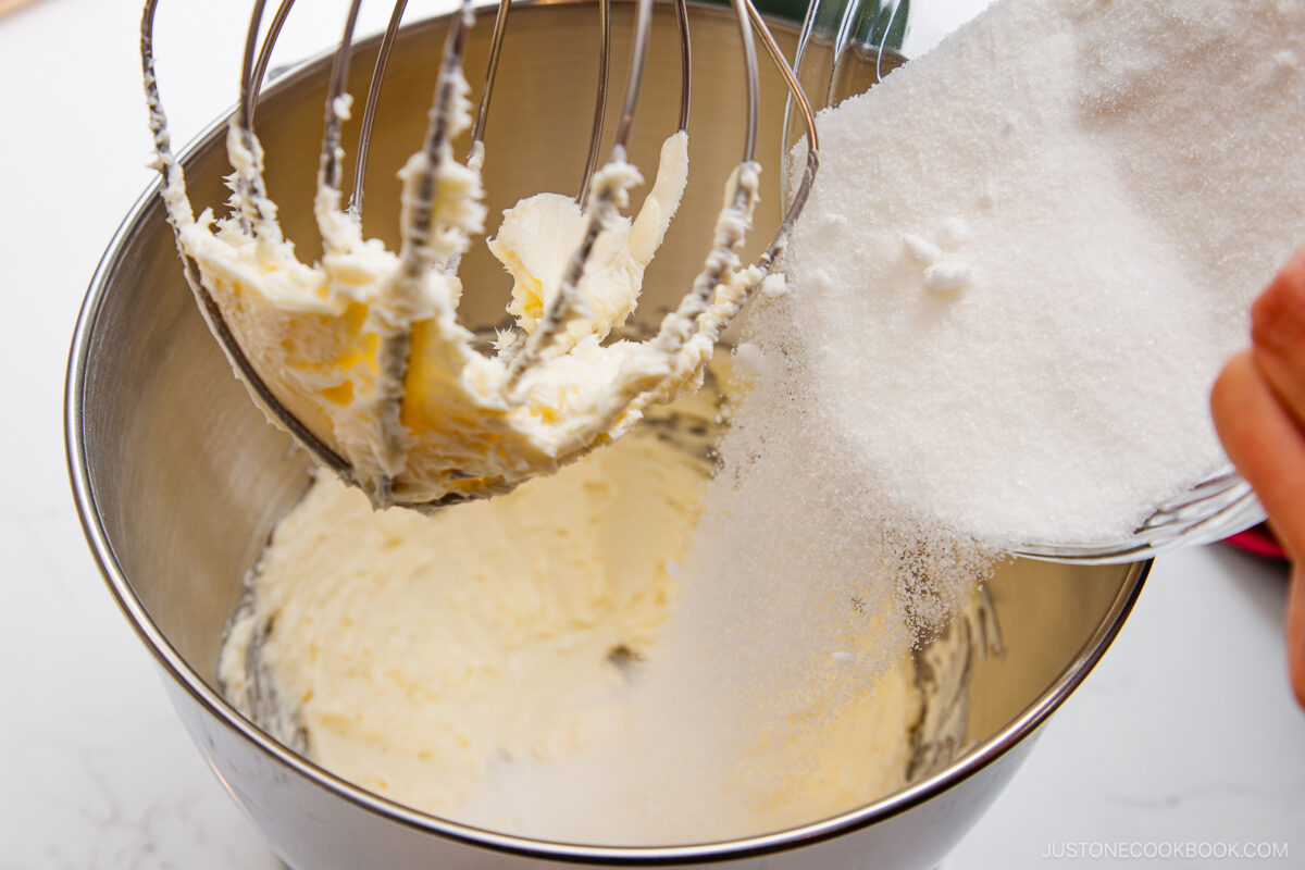A person pours granulated sugar from a bowl into a stand mixer with creamed butter, preparing a baking mixture.