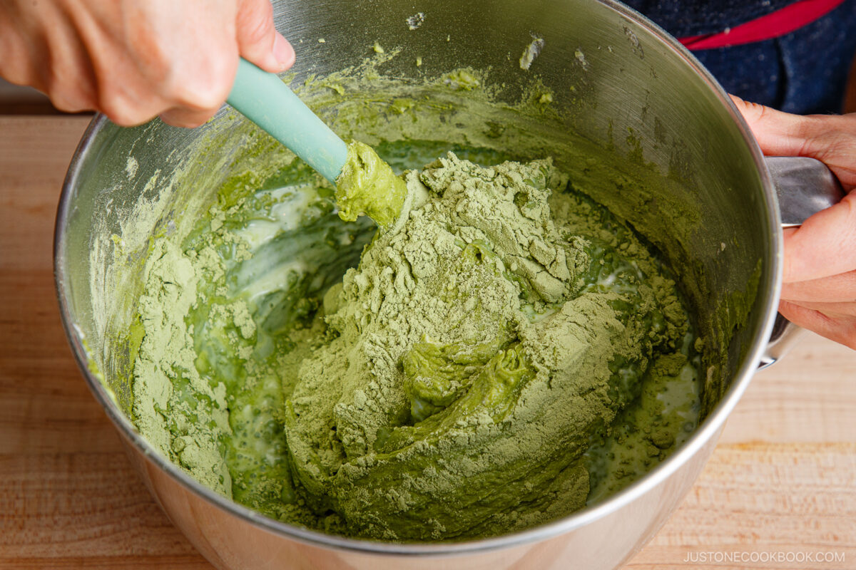 A person uses a spatula to mix thick green matcha batter in a large metal mixing bowl on a wooden surface.