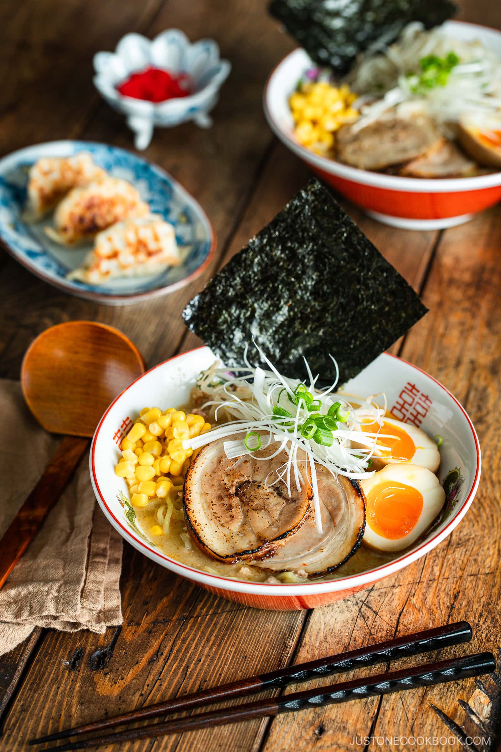 A bowl of ramen with sliced pork, soft-boiled eggs, corn, seaweed, and green onions sits on a wooden table next to chopsticks, a spoon, and a plate of dumplings in the background.